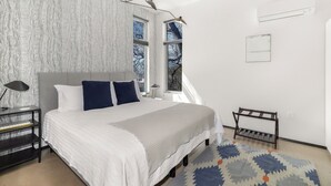 A bedroom with a large bed, two bedside lamps, a nightstand, a window with a view, and a wall-mounted air conditioner. - Sunny Chic Baca Railyard Retreat: Balcony + Full Kitchen + Laundry (Santa Fe)