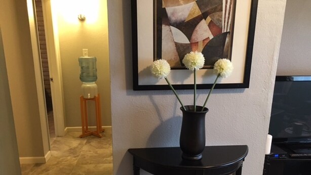 Un salon avec une table d’appoint noire sur laquelle est posé un vase rempli de fleurs blanches, un tableau accroché au mur et un distributeur d’eau en arrière-plan.
