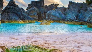 Una playa con agua turquesa cristalina, formaciones rocosas y una hoja de palmera en primer plano.