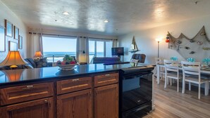A modern kitchen with wooden cabinets, a black stove, and a dining area with white chairs and a table. - Sands Lookout at Nye Beach - Beach front,Easy access condo with great ocean view (Newport)