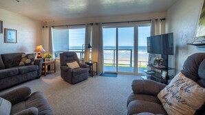 A spacious living room with a large window overlooking the beach, a comfortable sofa, a chair, and a TV on a stand. - Sands Lookout at Nye Beach - Beach front,Easy access condo with great ocean view (Newport)