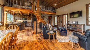 A spacious living area with a wooden floor, a staircase, a dining table with chairs, a sofa, a television, and a kitchen in the background.