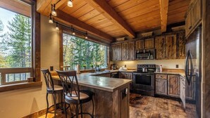 A rustic kitchen with wooden cabinets, a large island with bar stools, and a view of trees through the windows.