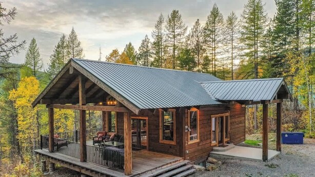 A wooden cabin with a metal roof, a porch with seating, and a gravel driveway.