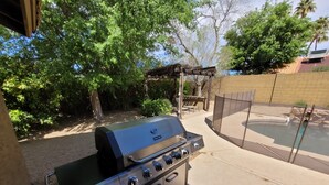 Een draagbare gasgrill met vier branders en een bijzettafeltje, in een achtertuin met een zwembad en een pergola.