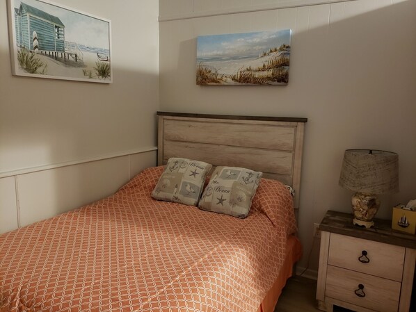 A bedroom with a wooden headboard, a patterned orange bedspread, two decorative pillows, a nightstand with a lamp, and two wall-mounted paintings.