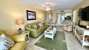 A living room with a green sofa, a yellow armchair, a dining table with chairs, a TV, and a kitchen in the background. - 7th Fl. Ocean Front Balcony💎So Clean, WIFI, Pools, Athletic Courts, Grills.. (Orange Beach)