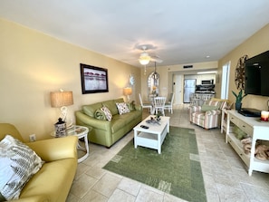 A living room with a green sofa, a yellow armchair, a dining table with chairs, a TV, and a kitchen in the background.