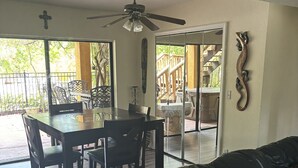 Un vaste espace de vie comprenant un canapé en cuir noir, une table à manger avec des chaises, un ventilateur de plafond et une vue sur une terrasse en bois, accessible par des portes vitrées.