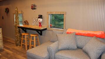 A cozy living room with a grey sofa, wooden stools, a small table with a snowman decoration, and a window with a view of greenery.