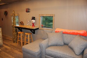 Ein gemütliches Wohnzimmer mit einem grauen Sofa, Holzschemeln, einem kleinen Tisch mit Schneemann-Dekoration und einem Fenster mit Blick auf Grünflächen.