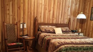 Una habitación con paneles de madera, una cama, un sillón mecedora, una mesita de noche con una lámpara y otra mesita con adornos.