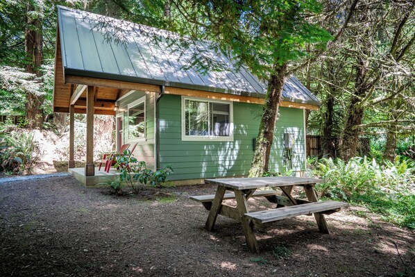 Una piccola baita di legno verde con tetto in metallo, un portico coperto e un tavolo da picnic in una zona boschiva.