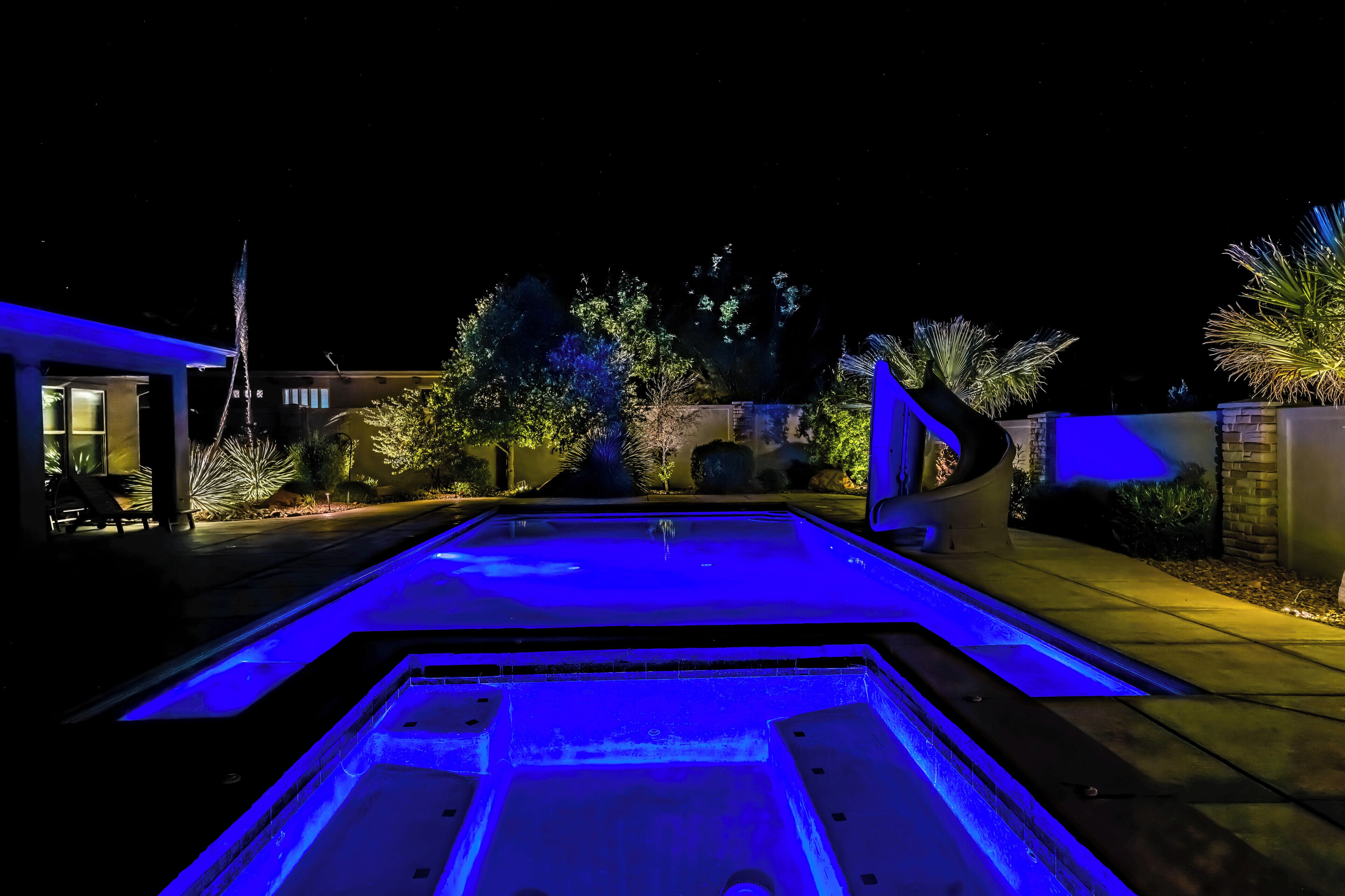 A swimming pool with blue lighting, surrounded by a landscaped garden and a building with a curved facade.
