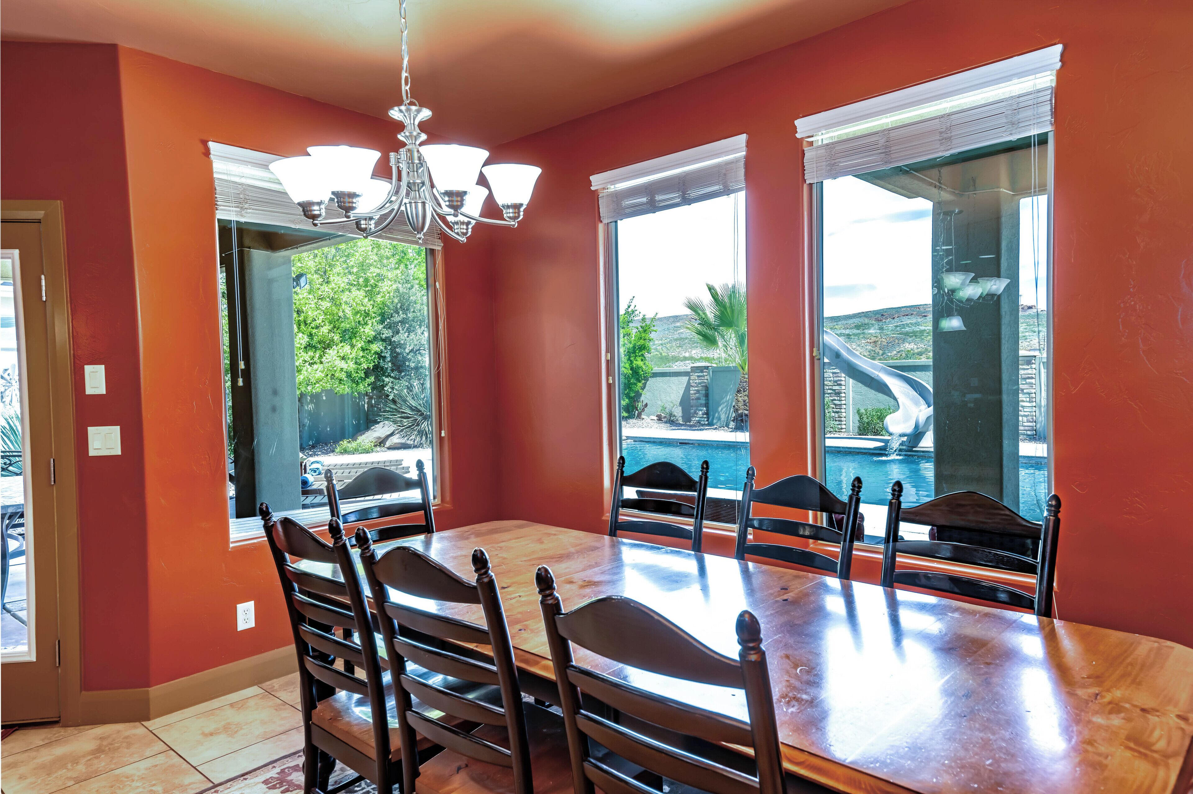 A dining area with a wooden table and chairs, large windows, and a chandelier.