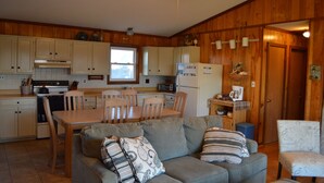 A compact kitchen with wooden cabinets, a dining table, and chairs. There is a gray sofa with patterned pillows in the living area. - Beach? We gotcha Beach-oceanside, only steps from uncrowded beautiful beach  (Nags Head)