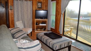 A cozy living room with a sofa, a television on a stand, and a sliding glass door leading to an outdoor deck. - Beach? We gotcha Beach-oceanside, only steps from uncrowded beautiful beach  (Nags Head)