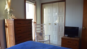 A bedroom with a bed, a wooden dresser, a white chair, a television on a wooden nightstand, and a white door. - Beach? We gotcha Beach-oceanside, only steps from uncrowded beautiful beach  (Nags Head)