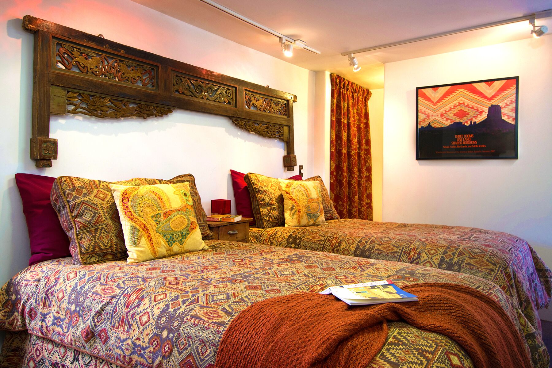 A bedroom with a patterned bedspread, multiple pillows, a wooden headboard, a red curtain, and a framed picture on the wall.