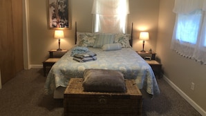 Un dormitorio con una cama, dos mesitas de noche con lámparas, un ventanal con cortinas traslúcidas, un cuadro en la pared y un cofre de mimbre.