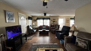 Una sala de estar con televisor, varias opciones de asiento, ventilador de techo y ventana con persianas.