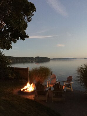 A lakeside view with a fire pit and Adirondack chairs.