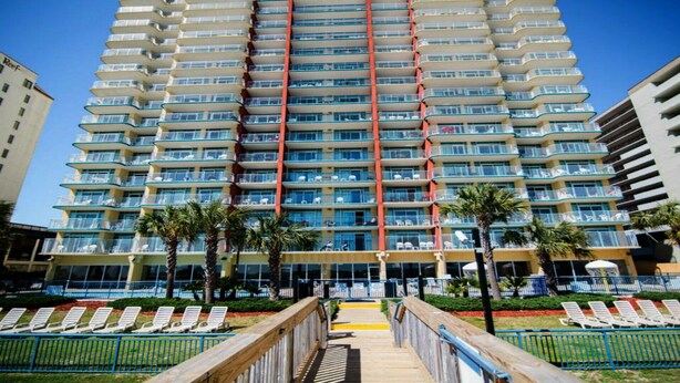 A multi-story hotel with a central red vertical feature, balconies, and a pool area with sun loungers. - Oceanfront Gem Clean 1080sf New Furniture, Beds, Carpet Free WiFi Cable Gym Park (Myrtle Beach)