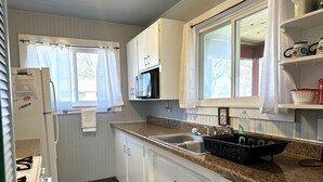 A compact kitchen with white cabinets, a microwave, a sink, and a window with curtains. - Clean, Family/Pet Friendly Lake View, 2-Bdrm Cabin Near Several Marinas/launches (Gamaliel)