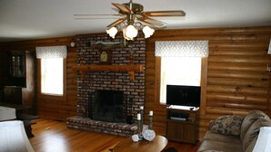 L’intérieur d’un chalet confortable, avec une cheminée, des murs en bois, un ventilateur de plafond et un téléviseur.