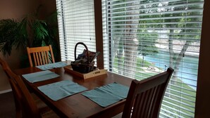 A dining area with a wooden table set for four, a wicker basket with magazines, and a view of a water body through large windows.