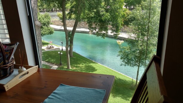 A view from a room with a wooden table and a window overlooking a river, a picnic table, and trees.