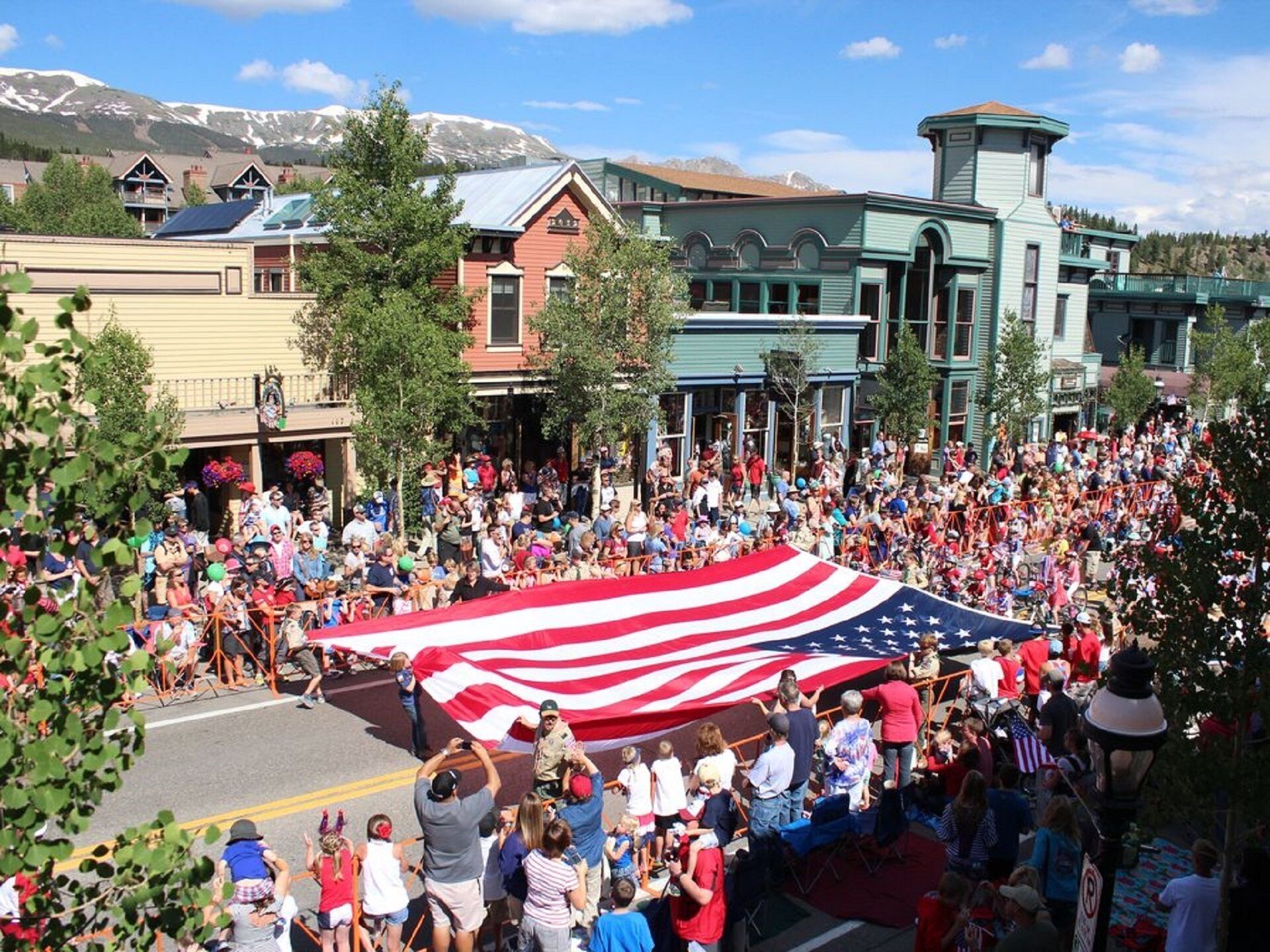 Ein Straßenumzug mit einer großen amerikanischen Flagge, umringt von Zuschauern und Gebäuden mit markanten Architekturstilen.