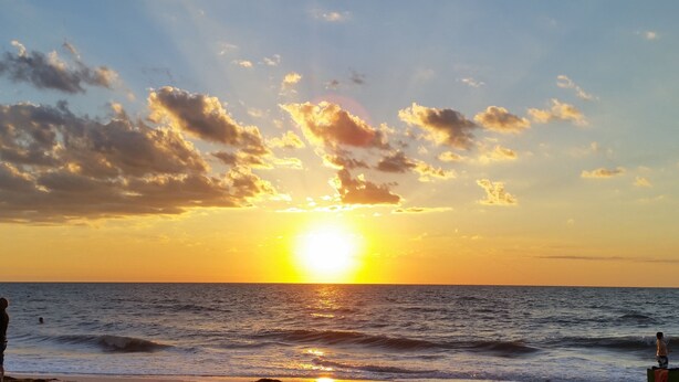 A sunset over the ocean with waves and a person standing on the shore.