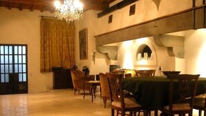 A dining area with a table set for four, surrounded by wooden chairs. A chandelier hangs from the ceiling, and a window with bars is visible on the left.