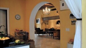 A kitchen with a sink, countertop, and a fruit bowl. There is a living area with a sofa and a chandelier. An archway connects the two spaces.