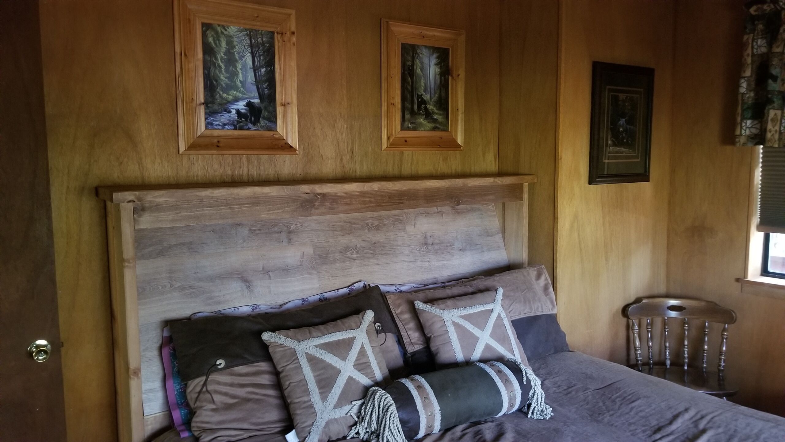 Un dormitorio con cabecera de madera, dos cuadros enmarcados, una cama con muchos cojines y un taburete de madera.