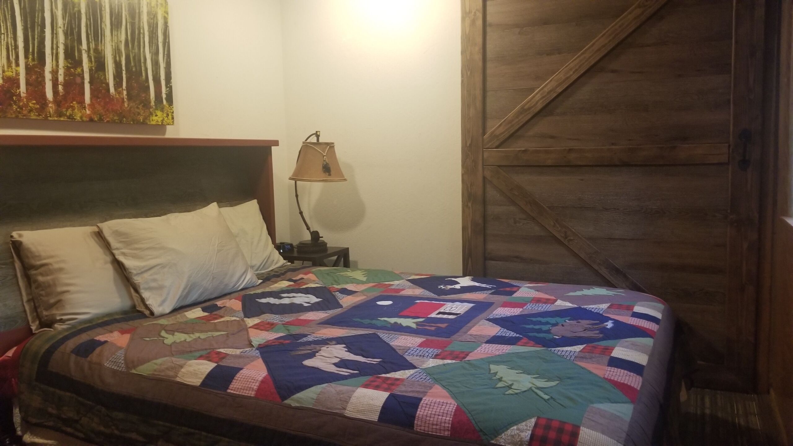 A bedroom with a wooden headboard, a quilted bedspread, a nightstand with a lamp, and a wooden door.