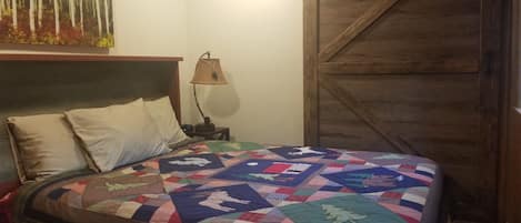 A bedroom with a wooden headboard, a quilted bedspread, a nightstand with a lamp, and a wooden door.
