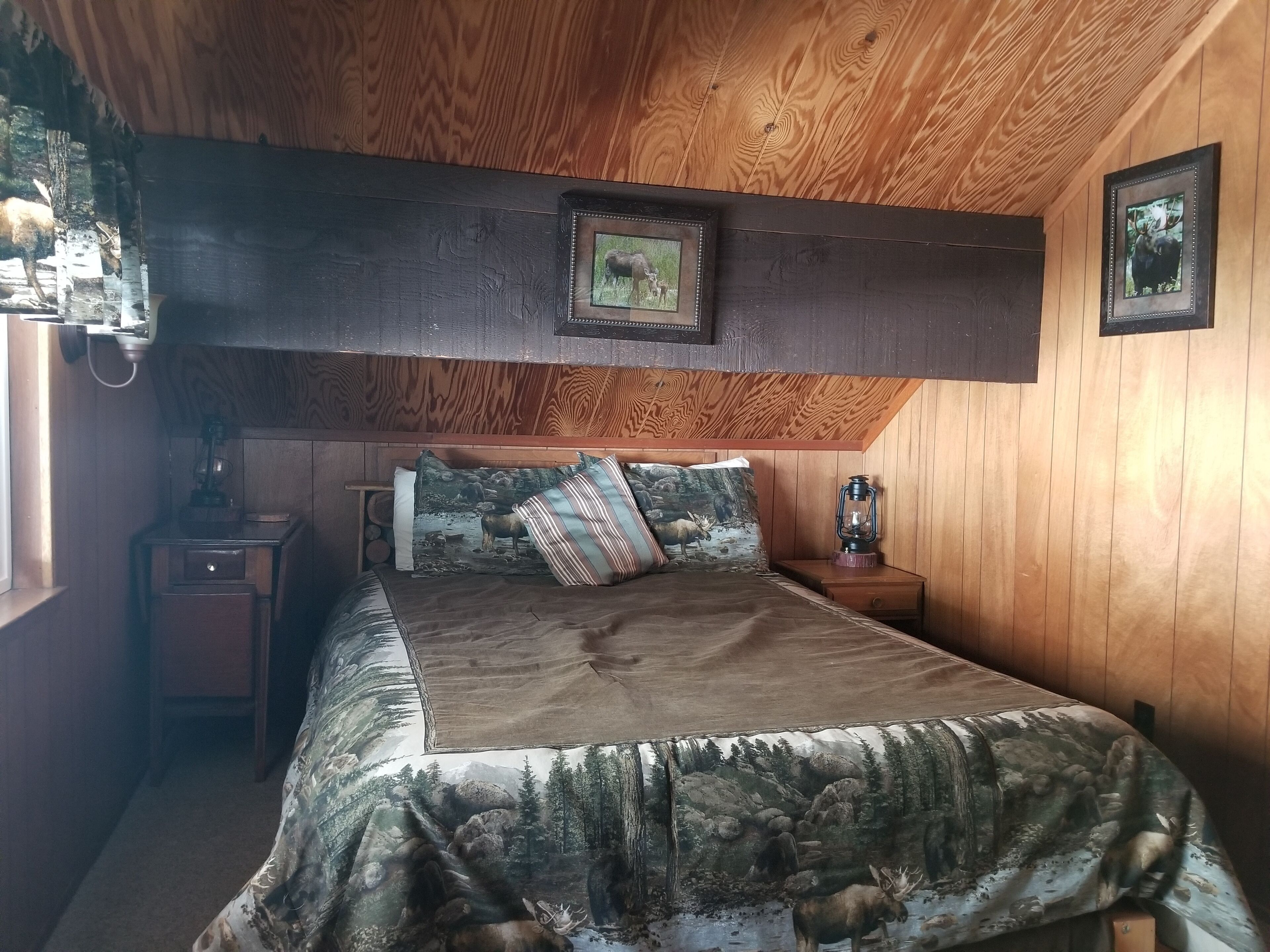 A bedroom with a wooden ceiling, a bed with a camouflage bedspread, two framed pictures on the wall, and a nightstand with a lantern.