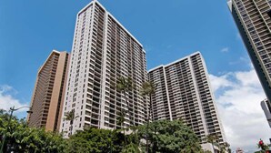 A high-rise building with a grid of windows and balconies, surrounded by greenery and other buildings.