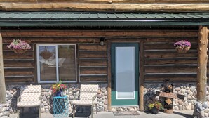 A log cabin with two chairs and a table outside, a stone wall, and a green door.