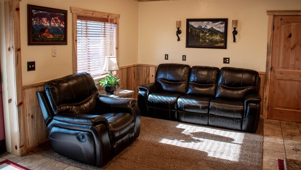 A living room with a black leather sofa, a recliner, and a wooden ceiling. There are two wall-mounted lamps and a framed picture on the wall.