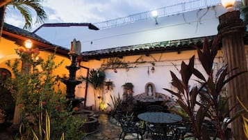 Une cour avec une fontaine en pierre, une table ronde et des chaises, le tout entouré de plantes et d’un mur décoratif.