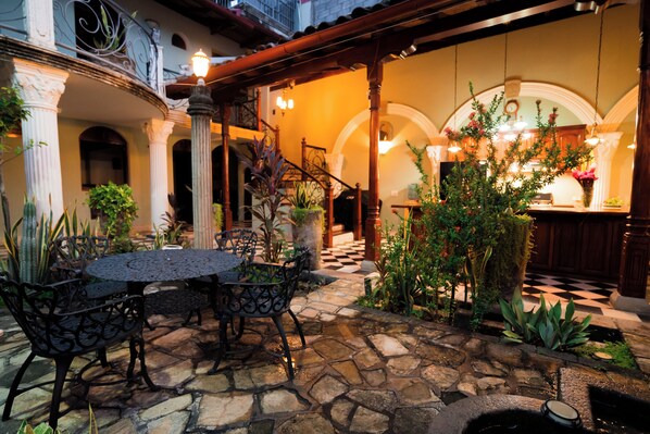 A stone-floored patio with a round table and chairs, surrounded by potted plants and a staircase leading to an upper level.