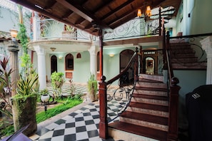 Una imponente escalera con barandales ornamentados que sube a un segundo piso, rodeada de abundante vegetación y un estampado de piso a cuadros.