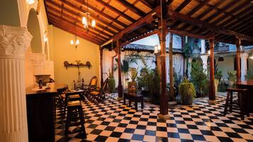 Un suelo a cuadros con techo de madera y columnas, una zona de bar con sillas y una mesa, y vistas a un jardín a través de un espacio abierto.