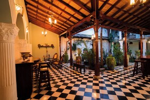 Un suelo a cuadros con techo de madera y columnas, una zona de bar con sillas y una mesa, y vistas a un jardín a través de un espacio abierto.