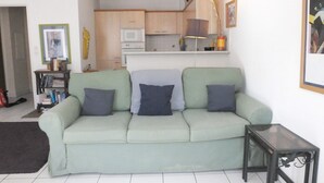 A living room with a green sofa, a small table, and a kitchen in the background.