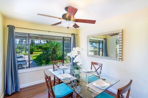 Un'area pranzo con tavolo in vetro, sedie, una ventola a soffitto, una finestra con tende e vista sul verde circostante.