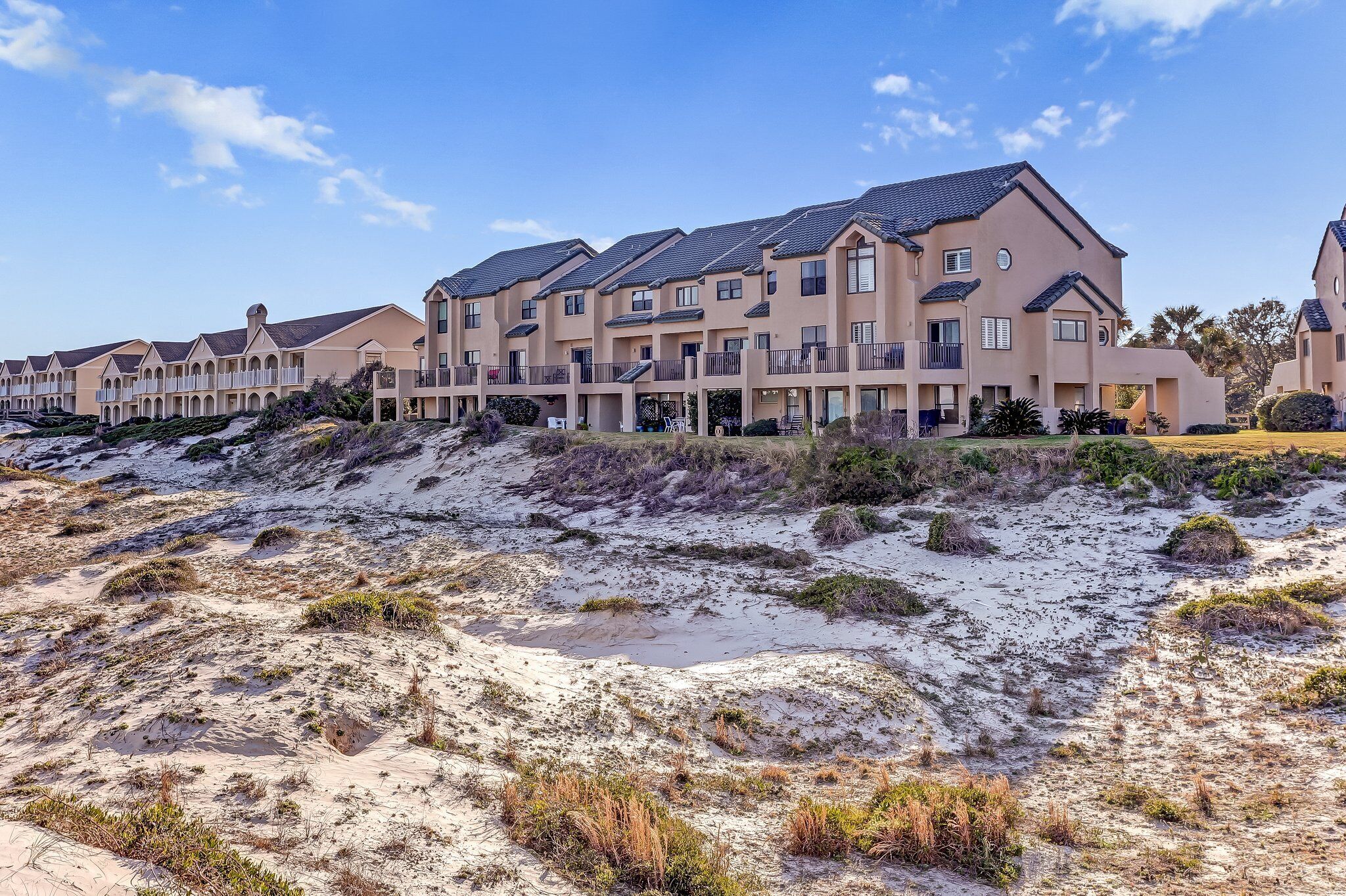 A row of modern multi-story buildings with balconies, situated on a sandy terrain with sparse vegetation.